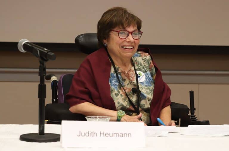 Disability advocate and pioneer Judith Heumann speaks at a conference. She is smiling, has red glasses on, is wearing a floral shirt, red shawl, and is sitting in her mechanical wheelchair.
