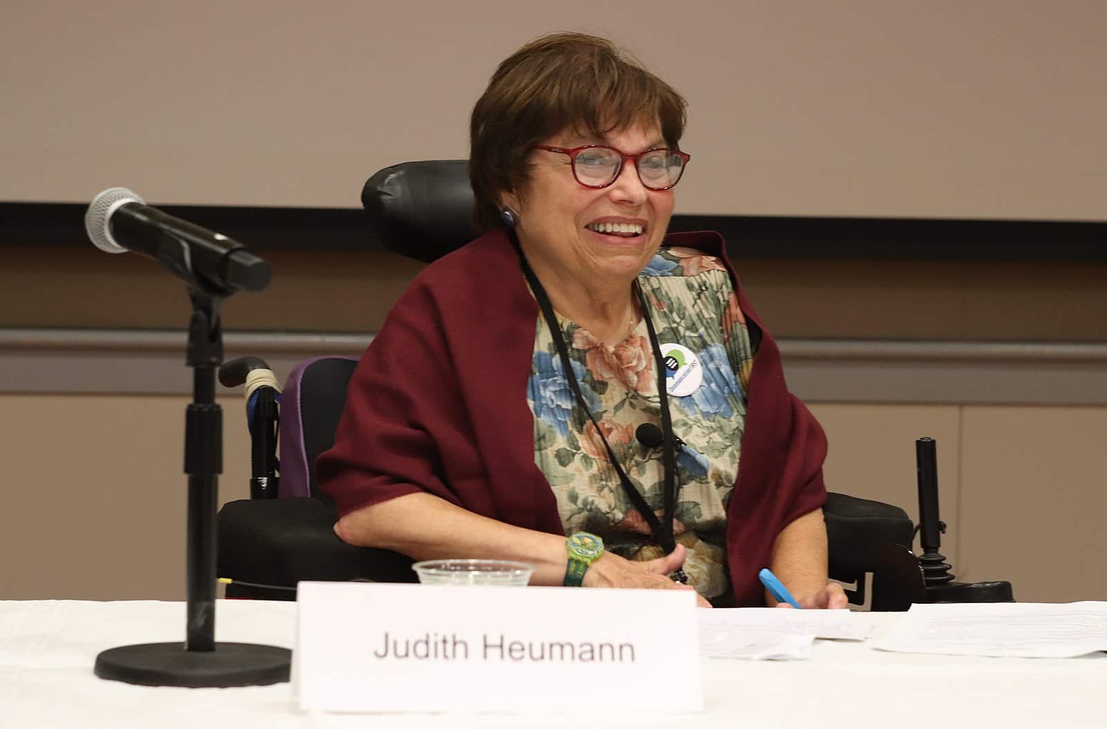 Disability advocate and pioneer Judith Heumann speaks at a conference. She is smiling, has red glasses on, is wearing a floral shirt, red shawl, and is sitting in her mechanical wheelchair.