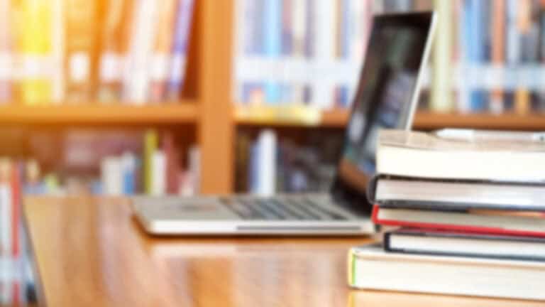 A desk in a college library, in the foreground is a stack of 5 textbooks, behind them is a silver laptop, in the background is a large book case with dozens of books.