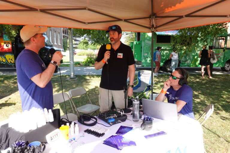 Marty Molloy, Mike Mangos, and Markus Goldman gather at a table covered in audio equipment under a canopy to record a podcast.