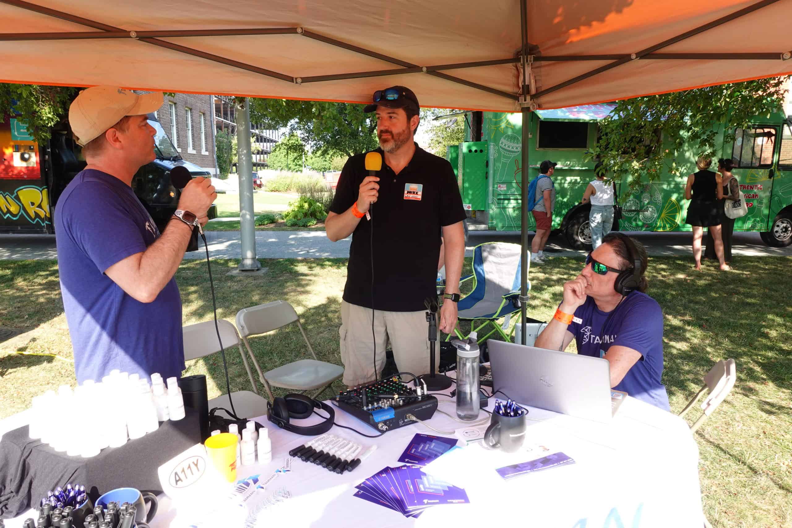 Marty Molloy, Mike Mangos, and Markus Goldman gather at a table covered in audio equipment under a canopy to record a podcast.