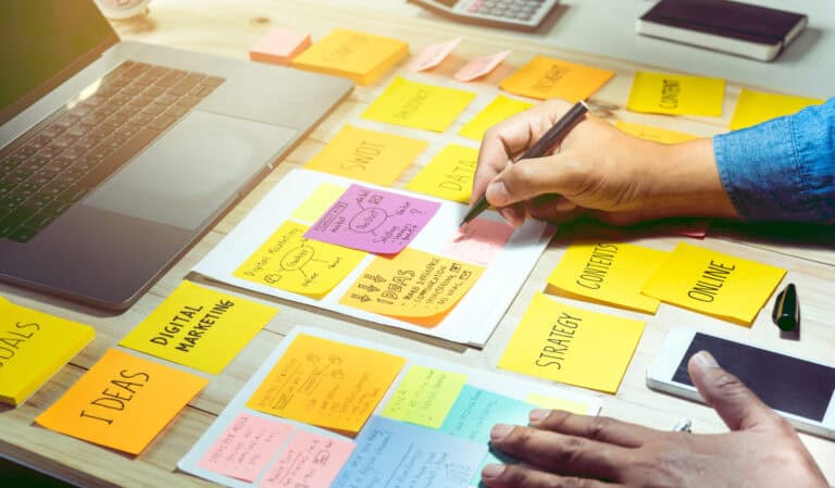 Digital marketing professional brainstorms his technical project goals on sticky notes spread across his desk. The sticky notes cover digital marketing strategy, communication ideas, online content, and other digital marketing needs.
