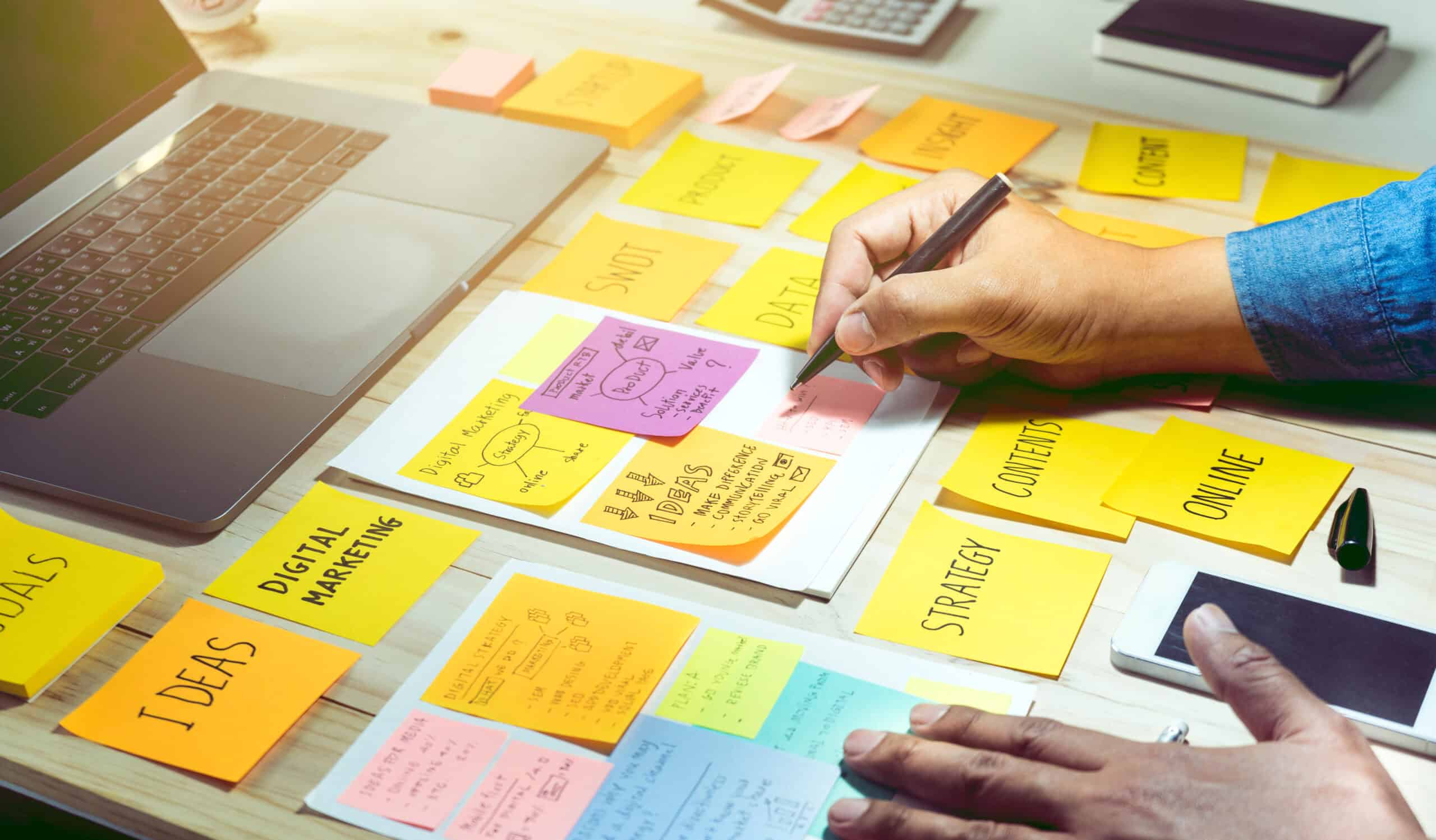 Digital marketing professional brainstorms his technical project goals on sticky notes spread across his desk. The sticky notes cover digital marketing strategy, communication ideas, online content, and other digital marketing needs.
