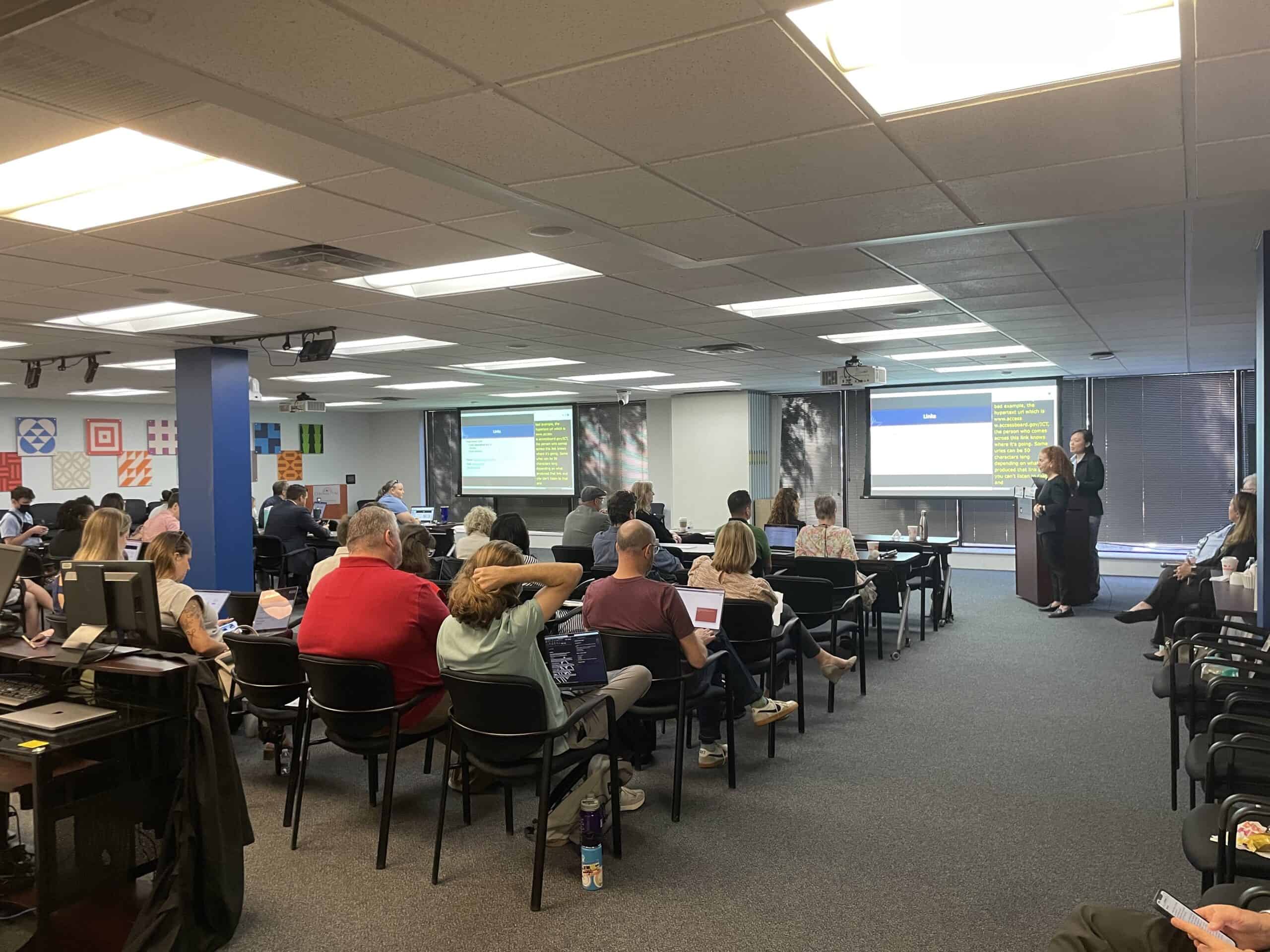A group of people attend a class on document accessibility. There is a presentation shown on two screens as an ASL interpreter translates for the instructor.