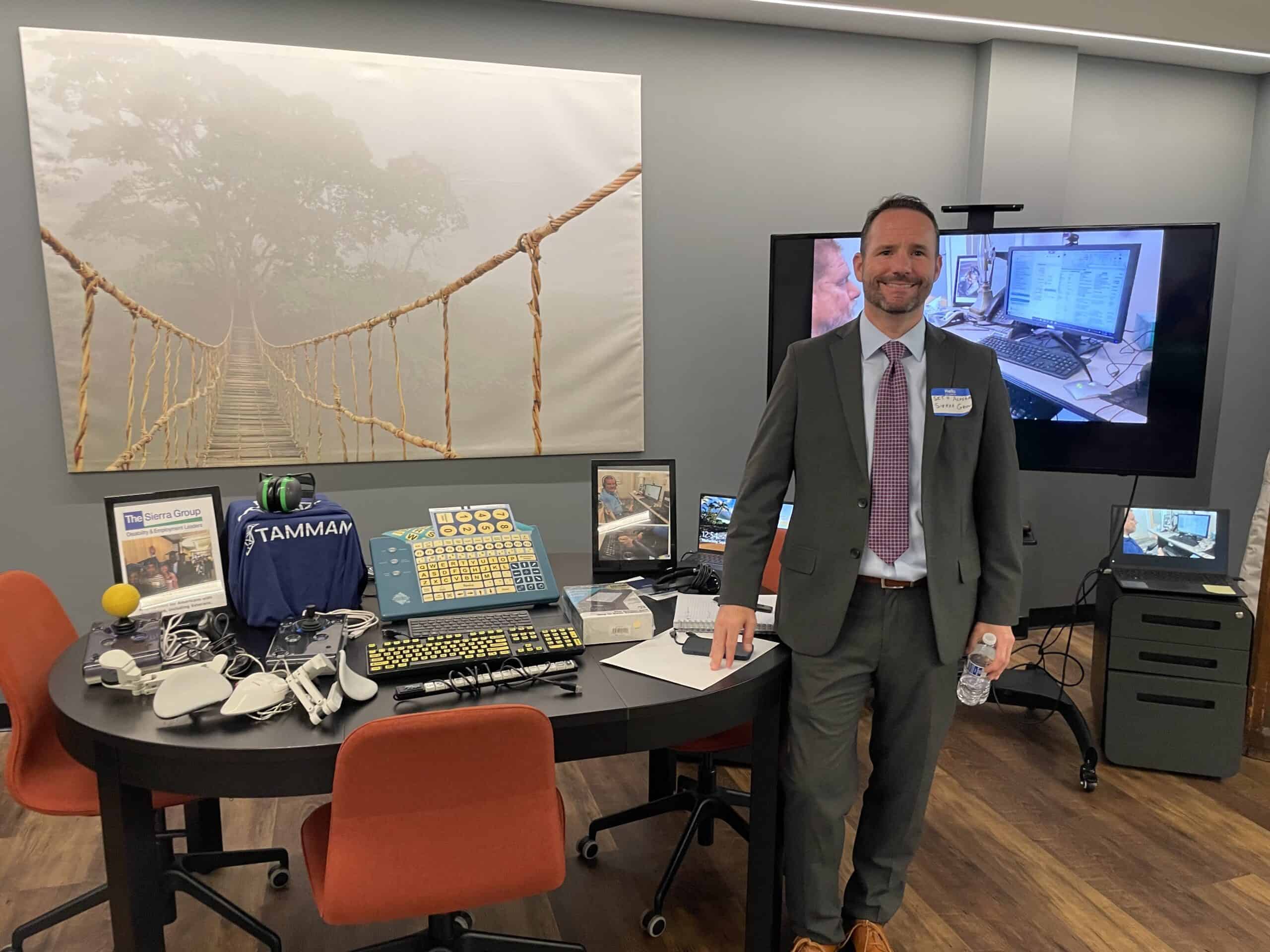 Seth Acosta from The Sierra Group stands near a table featuring many assistive devices, such as joy sticks, switches, high visibility keyboards and more.