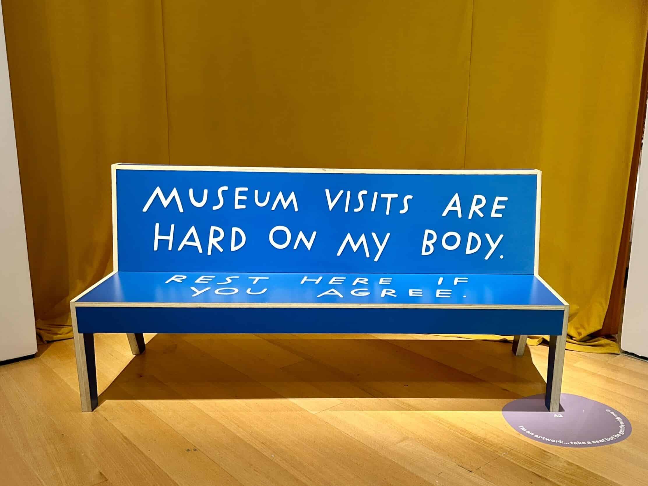 A six foot long wooden bench with straight edges colored blue with large capital letters that read: Museum visits are hard on my body rest here if you agree.