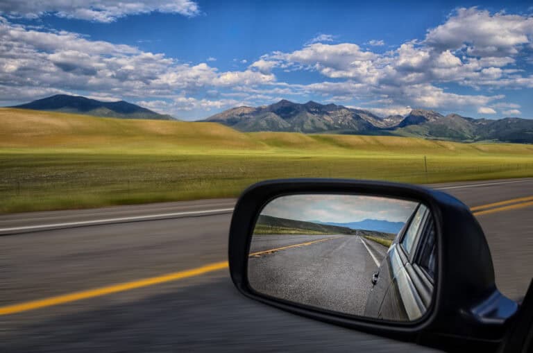 Side mirror of a vehicle with clear view of the road behind as the road ahead while larger view outside of mirror of grassy hills and mountains speed past in the distance