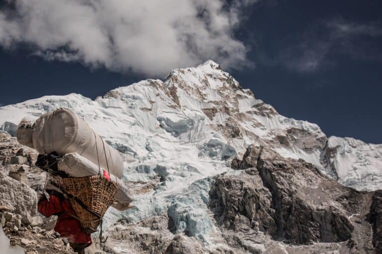 Sherpa with traditional basket carrying a load of supplies up a mountain with snow capped peak in distance
