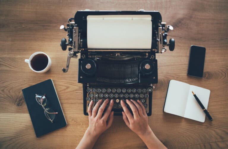 A writer uses a well-maintained typewriter. Next to them are glasses, black coffee, an open notebook with a pencil, and a smartphone.