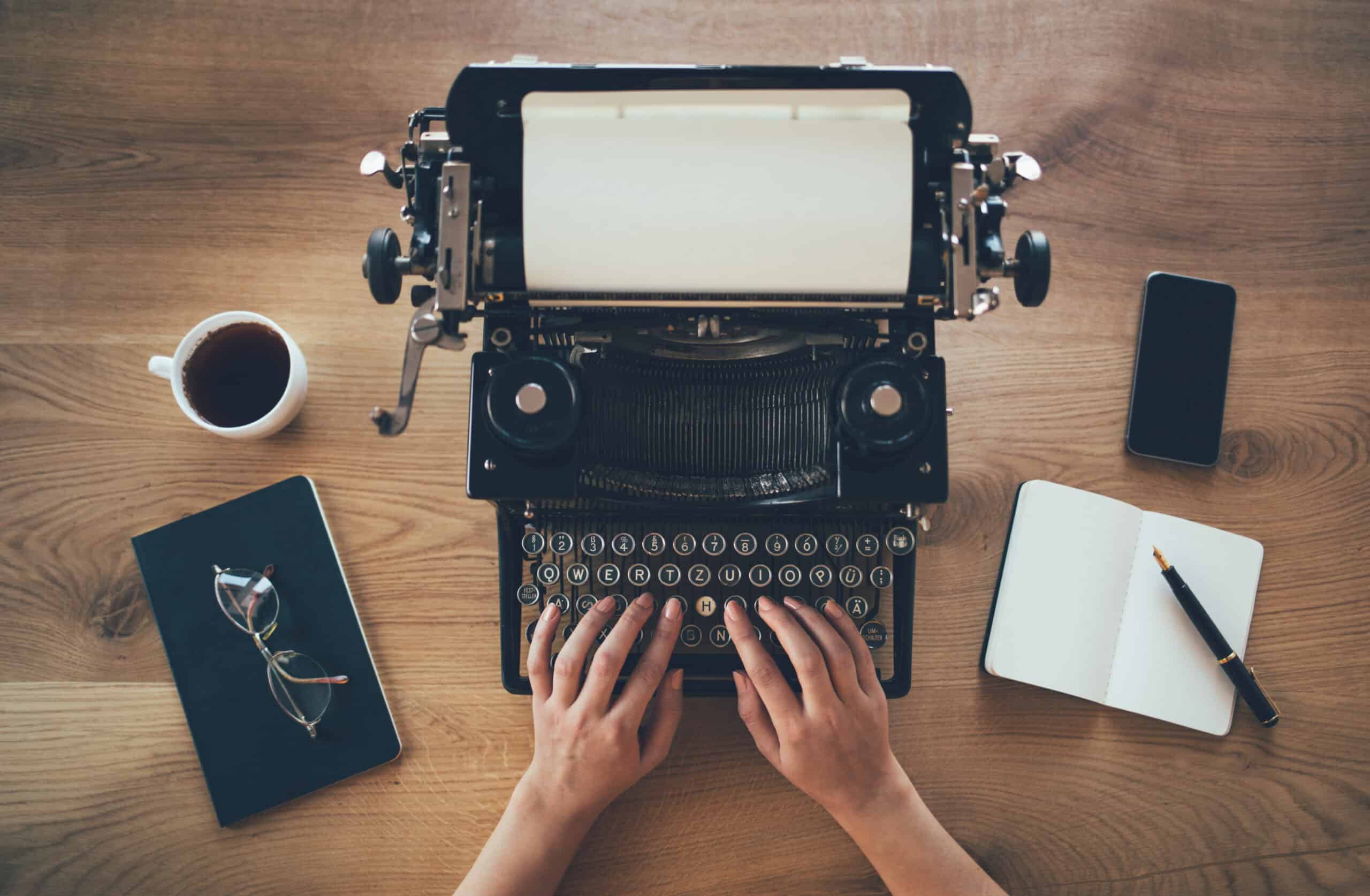 A writer uses a well-maintained typewriter. Next to them are glasses, black coffee, an open notebook with a pencil, and a smartphone.