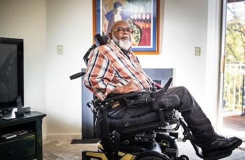 Theo Braddy sits in a power wheelchair with a smile on his face in a well-lit room. He is wearing glasses and a plaid shirt and sporting a beard.