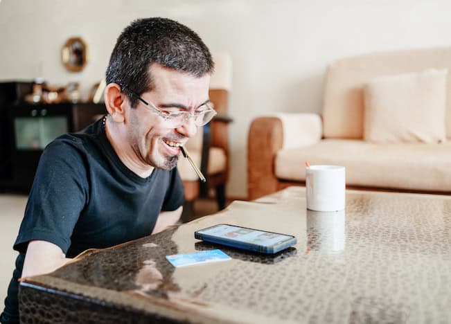 Person with physical use constraints using a stylus in mouth to navigate mobile device on table at home with credit card off to the side