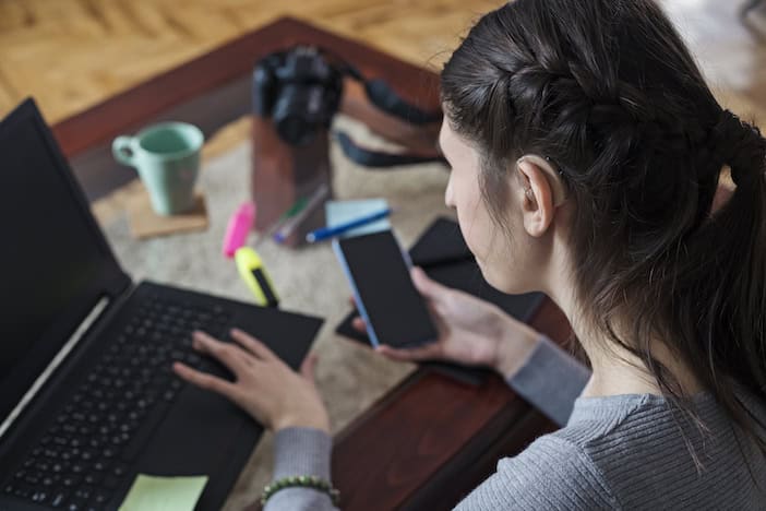 Person holding a smartphone in their right hand while using a laptop with their left hand on a a cluttered desk