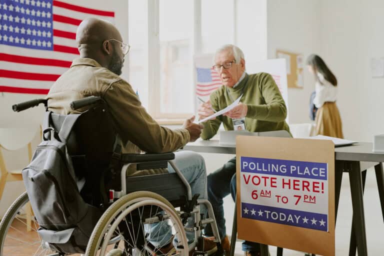 A man in a wheelchair is talking to a poll worker seated at a table inside a polling place. An American flag is displayed on the wall, and a sign on the table reads, 'Polling Place, Vote Here! 6 AM to 7 PM Today.'