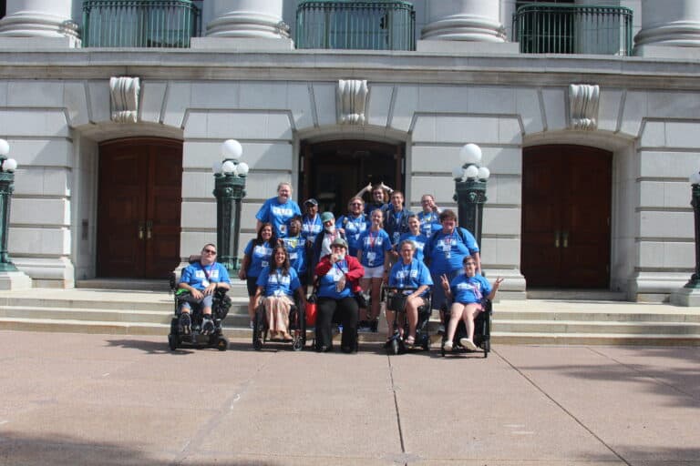 A group of people, including a number in wheelchairs, from the Wisconsin Youth Leadership Forum, wearing matching blue shirts smile in front of the Capitol Building in Madison.