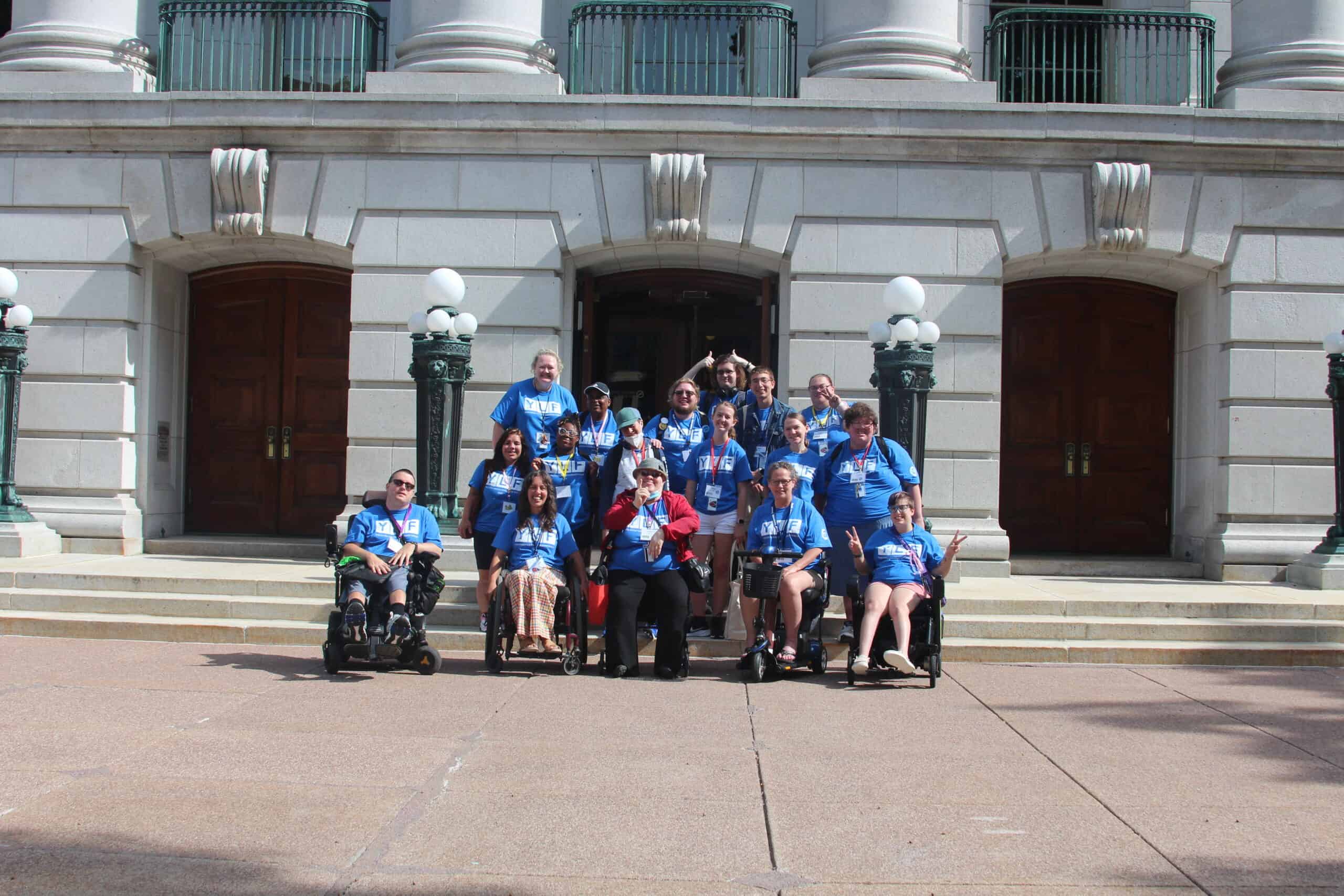 A group of people, including a number in wheelchairs, from the Wisconsin Youth Leadership Forum, wearing matching blue shirts smile in front of the Capitol Building in Madison.