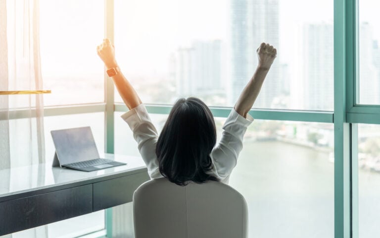 Person seated in front of high rise windows with back to camera and arms raised in a 'V' at desk with laptop