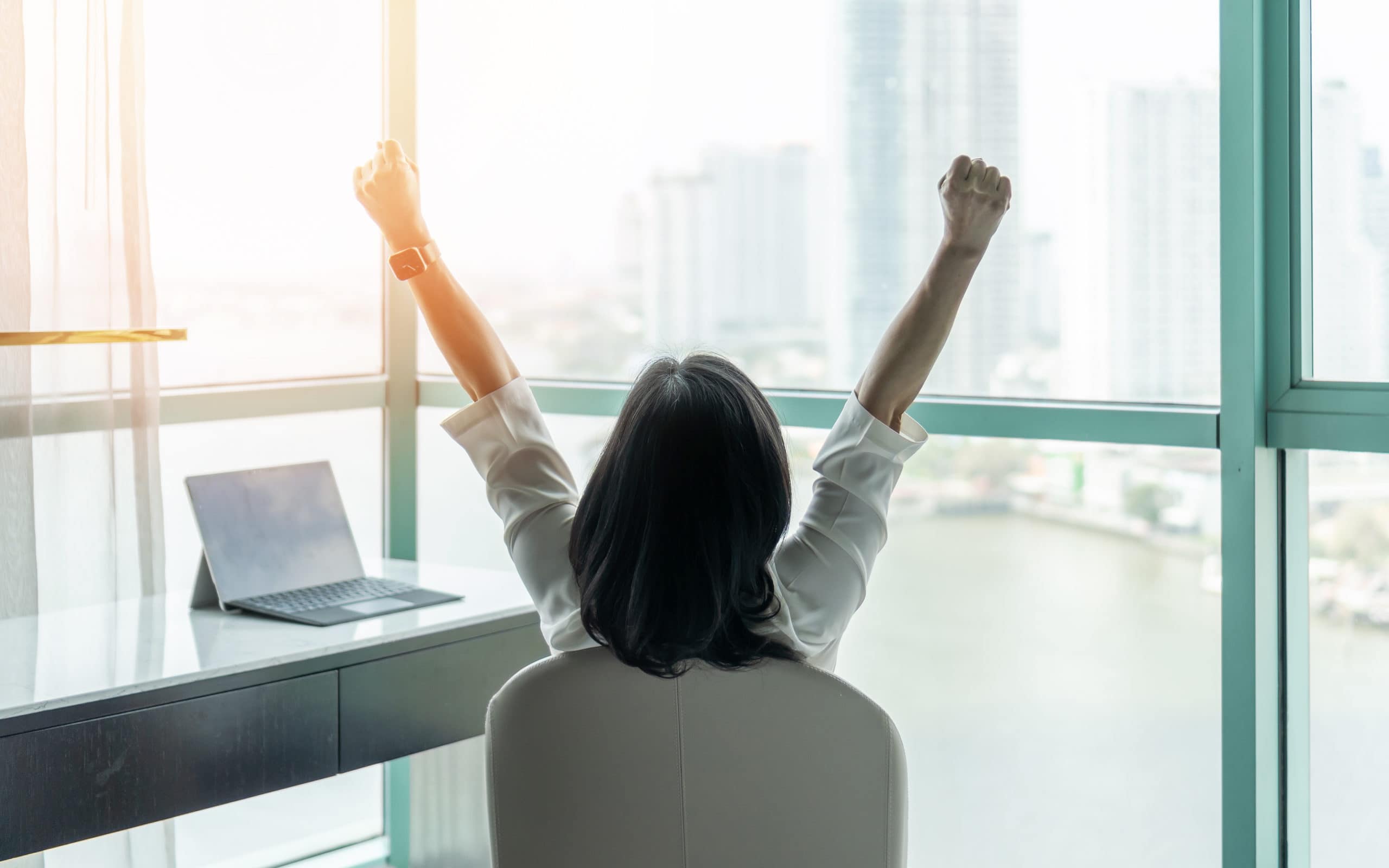 Person seated in front of high rise windows with back to camera and arms raised in a 'V' at desk with laptop