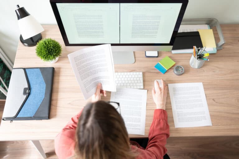 A digital document remediation subject matter expert changes a document to be more accessible to someone at her desk. In her hand is a printed copy of the document she is remediating on her computer.