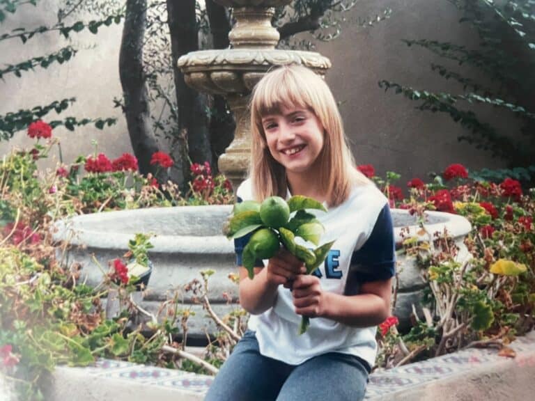 Raquella Freeman as a young girl. She is smiling at the camera while sitting on a stone fountain holding a bunch of green limes.
