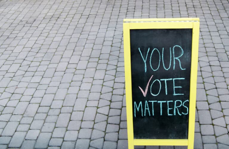 An A-frame chalkboard sign reads 'YOUR VOTE MATTERS,' with a checkmark next to the word 'VOTE. The yellow-framed sign is situated on a paved walkway.
