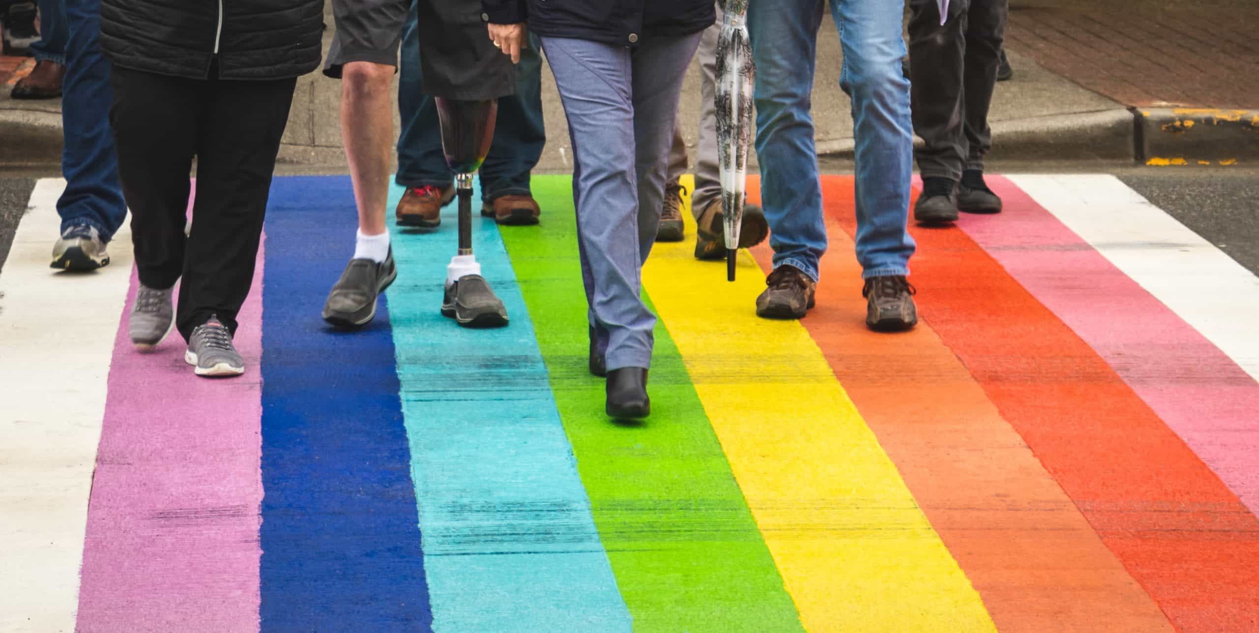 Eight people beginning to cross a rainbow flag painted street all depicted with legs only from the hip down and one person with a prosthetic left leg