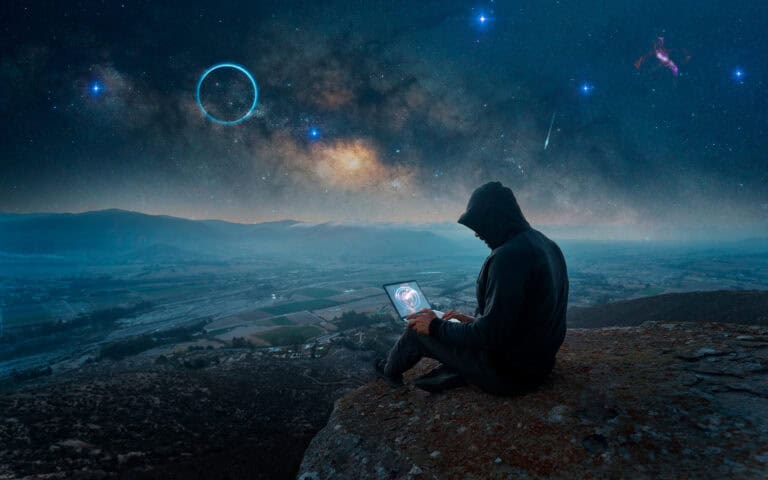 Hooded man with laptop on a rocky outcropping with night sky filled with galaxy of bright stars and planets