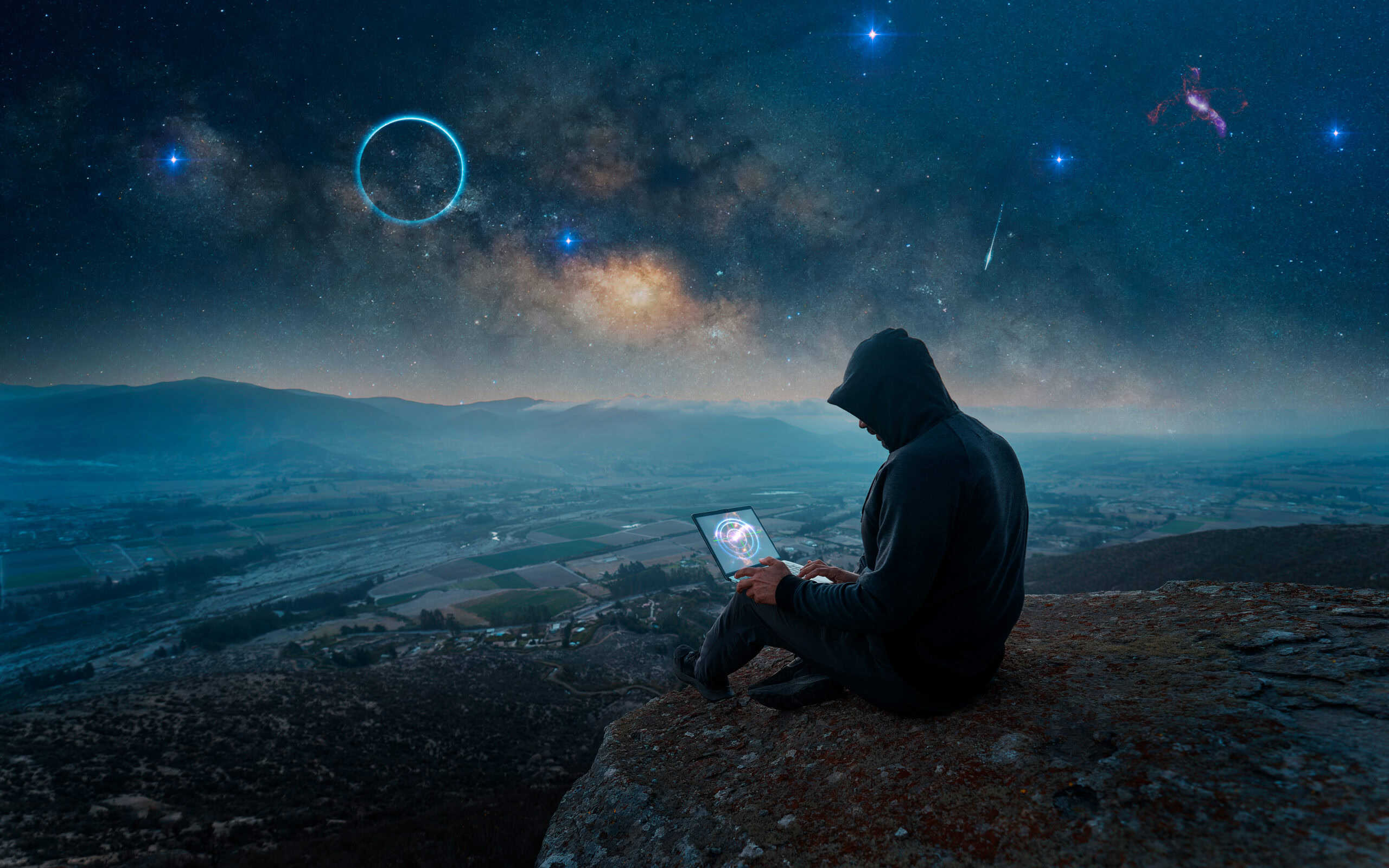 Hooded man with laptop on a rocky outcropping with night sky filled with galaxy of bright stars and planets