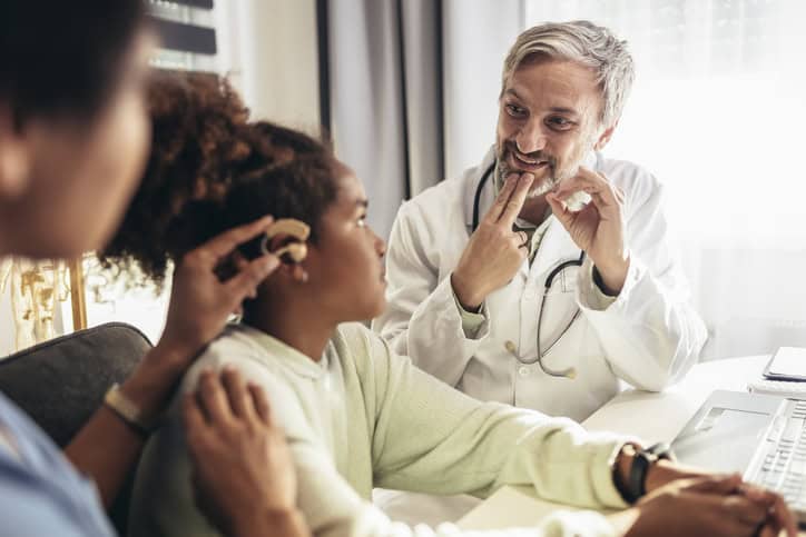 A young girl with a hearing aid sits in a doctor's office, watching a male doctor with gray hair and a smile as he communicates using sign language.