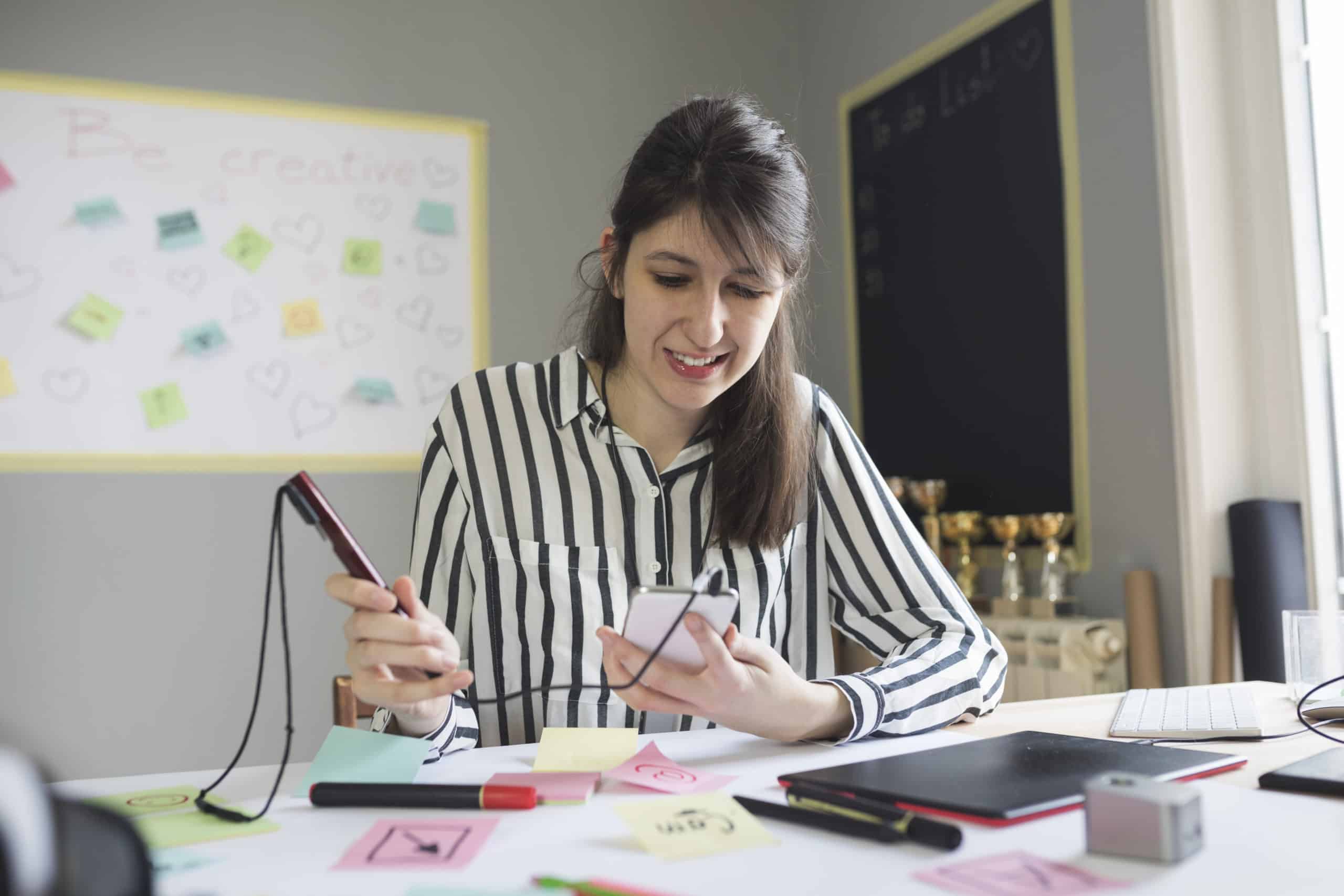 Designer with hearing aid seated at post-it strewn desk, using a recording pen hooked up to smart phone with tablet nearby