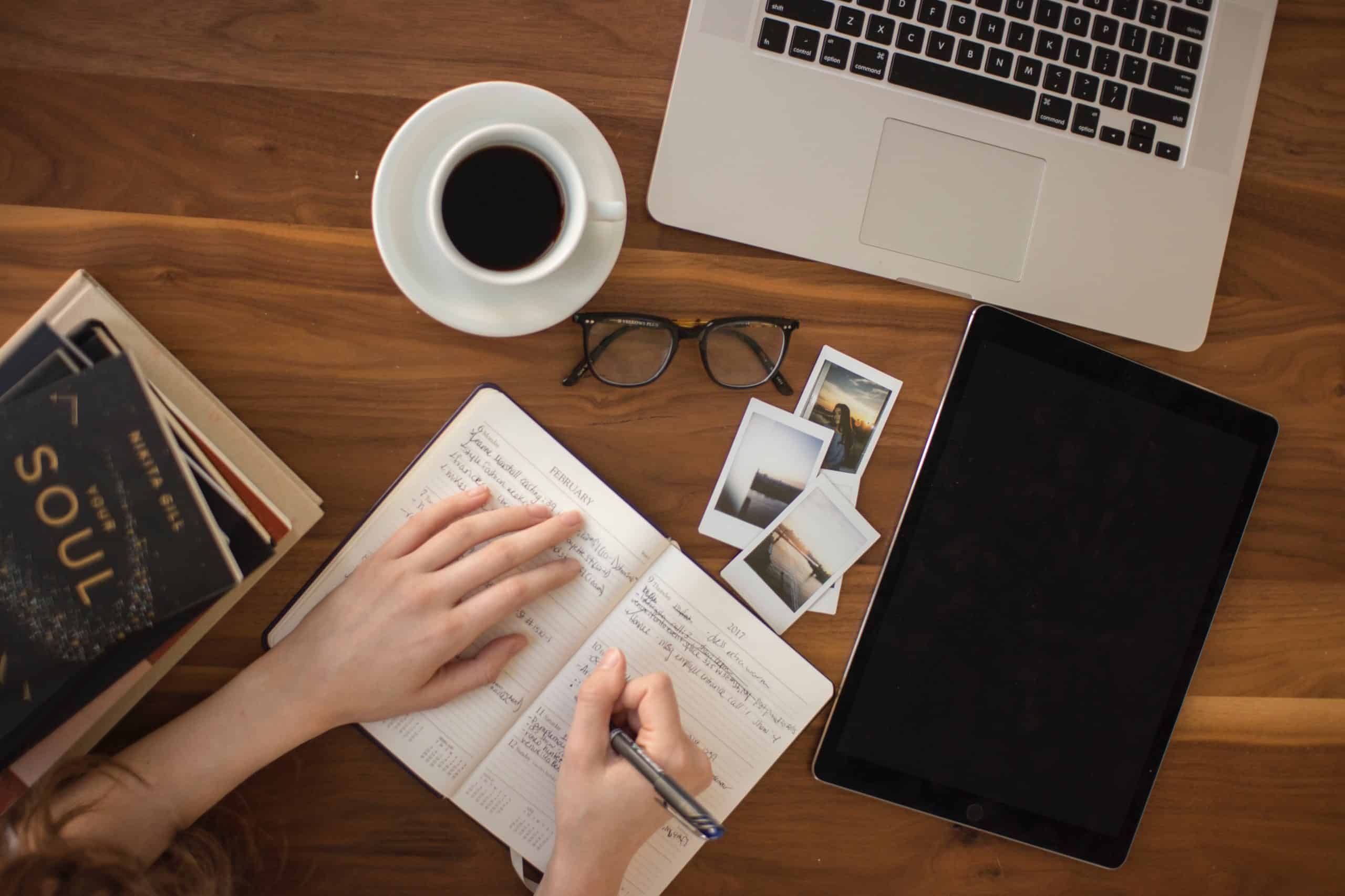 Aerial view of person writing in journal with a stack of books to the left, a coffee filled mug, glasses Polaroids, iPad and laptop arrayed on desk