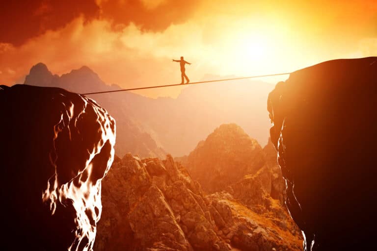A person tightrope walking between two cliffs in front of a sunset in a mountainous landscape
