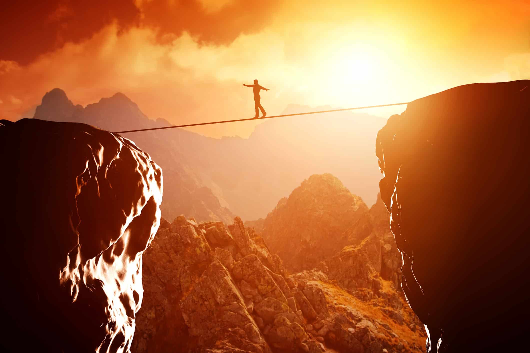 A person tightrope walking between two cliffs in front of a sunset in a mountainous landscape